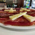 Tabla de embutidos ibéricos con lonchas de jamón, lomo y salchichón, acompañada de triángulos de queso curado con aceite de oliva.