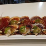 Zamburiñas a la plancha servidas en su concha con salsa verde y tiras de guindilla, acompañadas de un limón decorado.