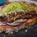 Tosta de carne a la parrilla con lechuga, queso rallado, cebolla morada, tomate y salsa, servida sobre pan artesanal.