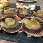 Zamburiñas a la plancha con salsa verde servidas sobre una base de pizarra, acompañadas de limón y salsas.