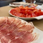 Plato de jamón curado acompañado de tomate fresco y una copa de sidra en una mesa de bar.