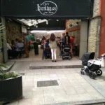 Entrada del bar-tienda Lantigua con varias personas disfrutando en la terraza y un hombre con un carrito de bebé.