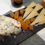 Tabla de quesos con cuñas de queso curado, queso azul y cubos de membrillo, acompañada de pan.